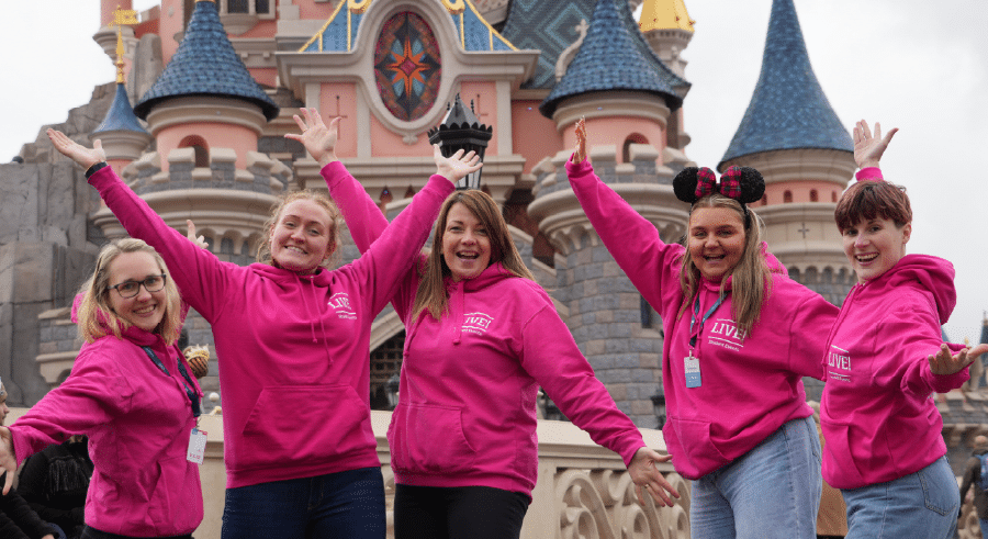 Five people in matching pink hoodies pose with arms raised and smiling in front of a fairytale castle at an amusement park.