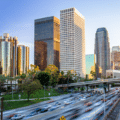 City centre cityscape with tall office buildings and palm trees, overlooking a busy multi-lane motorway with cars in motion at sunset.