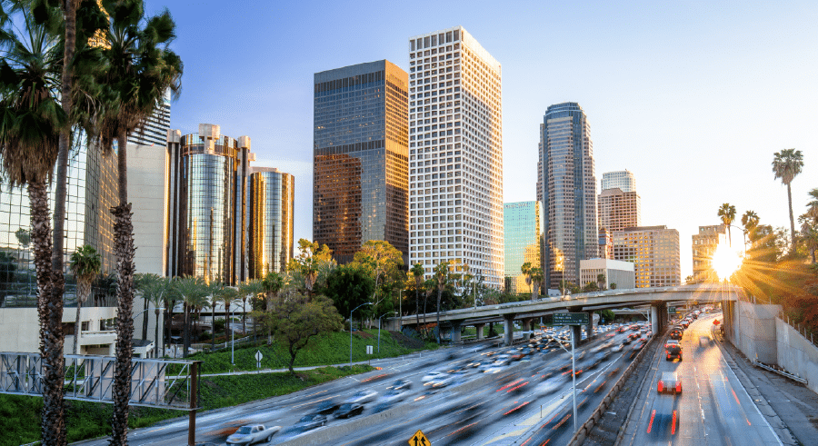 City centre cityscape with tall office buildings, palm trees, and a busy motorway with cars in motion during sunset.