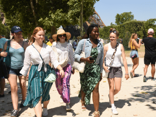 A group of young women walk together outdoors on a sunny day, with trees and other people in the background.
