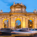 Cars pass in front of the illuminated Puerta de Alcalá, a neoclassical monument with arches and statues, at dusk in Madrid, Spain.