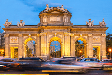 Cars pass in front of the illuminated Puerta de Alcalá, a neoclassical monument with arches and statues, at dusk in Madrid, Spain.