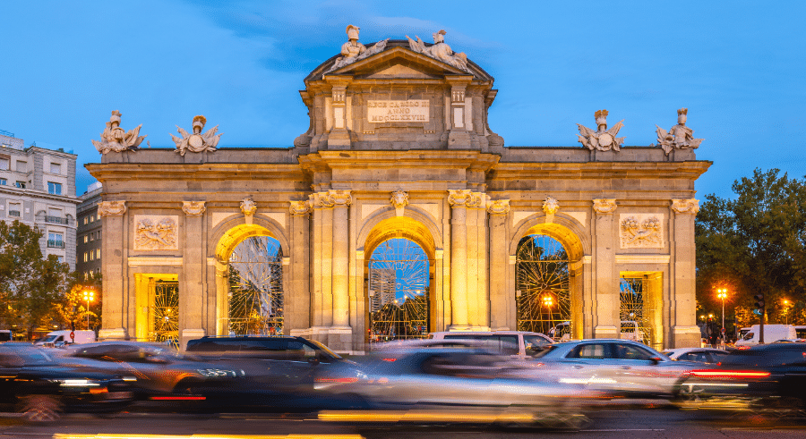 Stone gate with three large arches and two smaller side arches, illuminated at twilight, with cars passing in front and trees in the background.