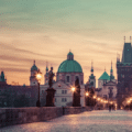 Charles Bridge in Prague at dawn, featuring historic statues, street lamps, and the city skyline with domes and towers under a pastel sky.