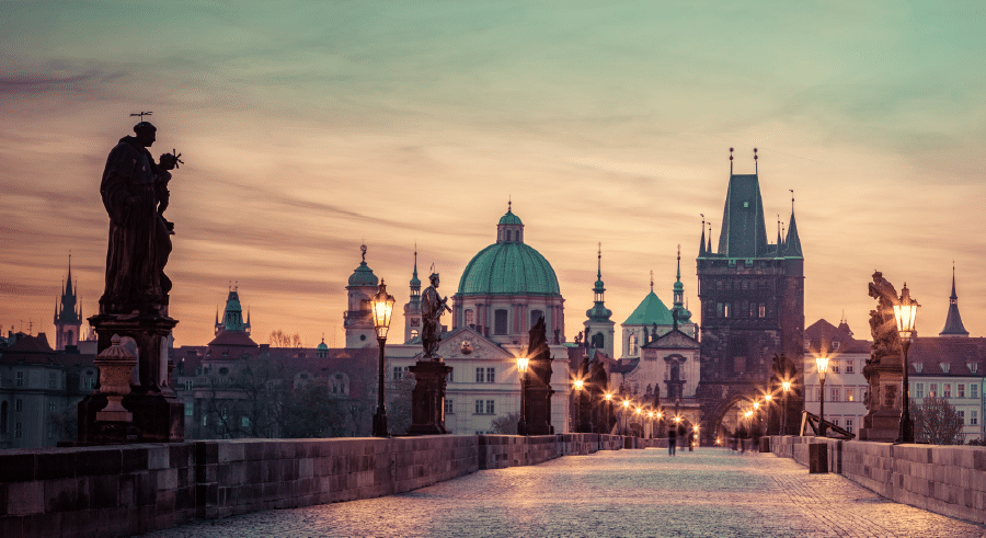 Charles Bridge in Prague at dawn, featuring historic statues, street lamps, and the city skyline with domes and towers under a pastel sky.