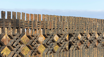 A close-up view of a wooden fence with an intricate geometric pattern, set against a blurred outdoor background under a clear sky.