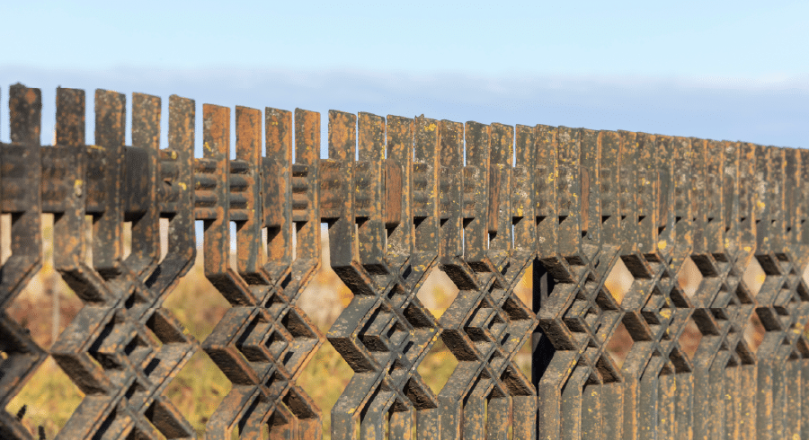 Close-up of a decorative metal fence with geometric patterns, showing signs of rust and weathering, with blurred grass and sky in the background.