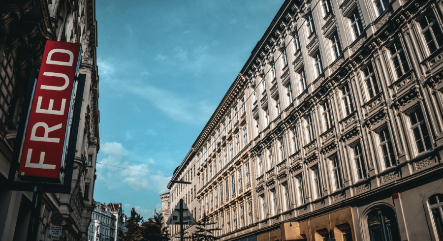 A city street with ornate historic buildings and a large red sign reading "FREUD" on the left side, under a partly cloudy blue sky.