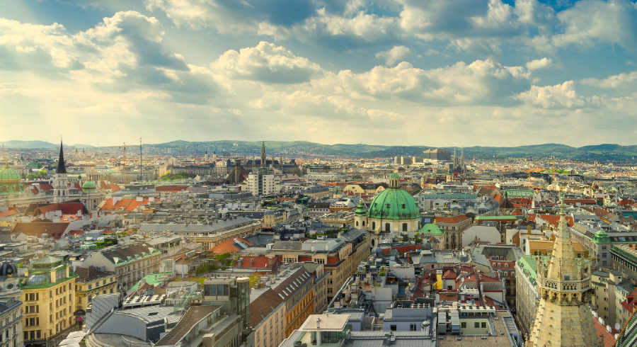 A panoramic view of a European city with historic buildings, a green-domed structure, church spires, and a hilly landscape under a partly cloudy sky.