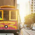 A red and yellow San Francisco cable car travels on rails down a city street lined with buildings and cars.