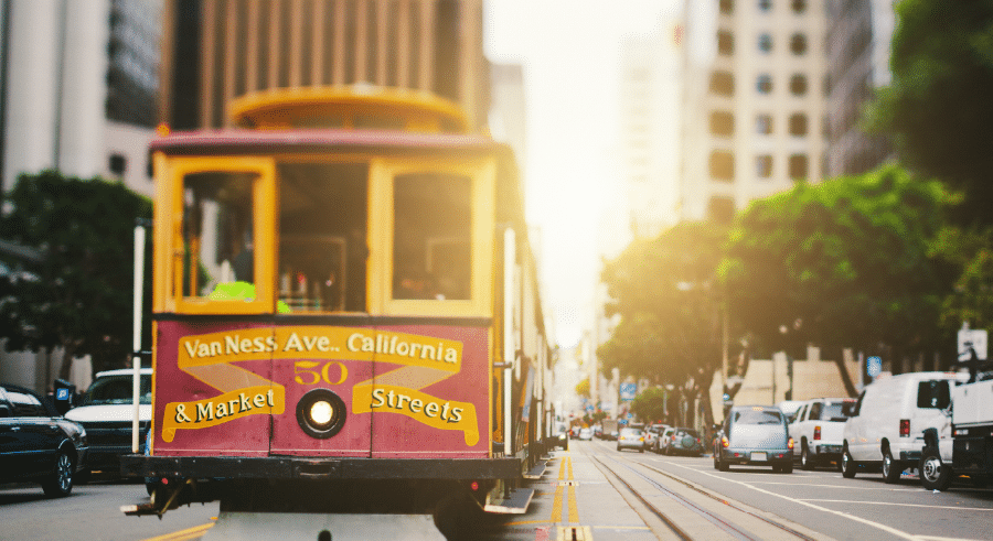 San Francisco cable car marked "Van Ness Ave. California & Market Streets" travels along city tracks with tall buildings and parked cars in the background.