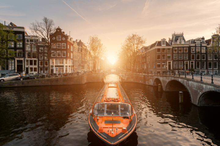 A canal boat travels under a bridge in Amsterdam at sunset, surrounded by traditional Dutch buildings and trees lining the waterway.