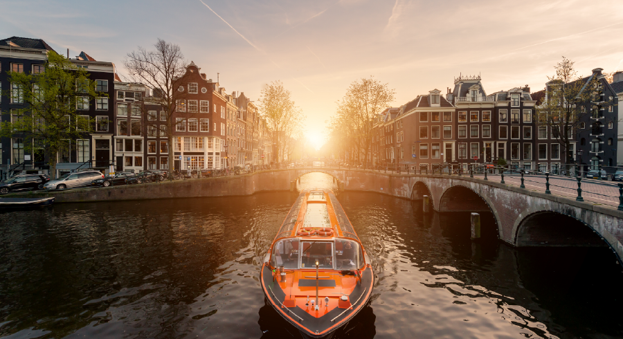 A canal boat travels under a bridge in Amsterdam at sunset, surrounded by traditional Dutch buildings and trees lining the waterway.