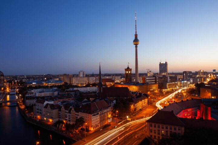 A cityscape of Berlin at dusk featuring the Fernsehturm TV tower, illuminated streets, and buildings under a clear evening sky.