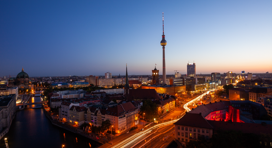 A cityscape of Berlin at dusk featuring the Fernsehturm TV tower, illuminated streets, and buildings under a clear evening sky.