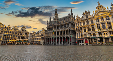 A large cobblestone square surrounded by ornate historic buildings with elaborate façades at sunset.