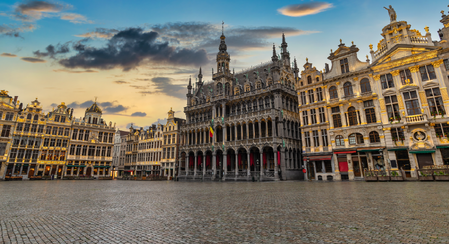 The image shows the Grand Place in Brussels at sunset, featuring ornate historic buildings and a cobbled square under a partly cloudy sky.