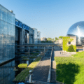 Modern glass buildings and a large reflective dome are seen near water and greenery on a clear, sunny day.