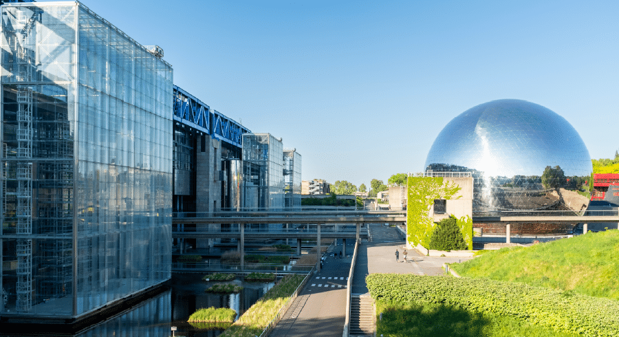 Modern glass buildings and a large silver sphere are shown on a bright, clear day, with pathways and greenery between the structures.