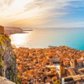 A coastal town with terracotta rooftops stretches to the sea under a sunset sky, viewed from a clifftop vantage point.