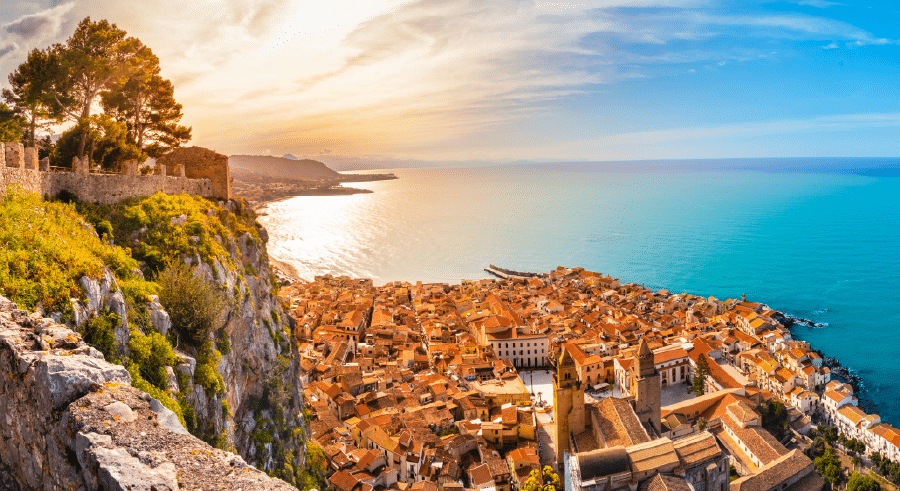A coastal town with orange-roofed buildings sits beside a bright blue sea under a sunny sky, viewed from a high stone wall surrounded by greenery.
