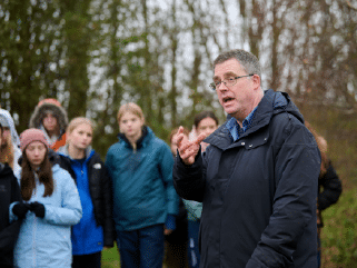 A man in a dark jacket speaks outdoors to a group of children and teenagers standing in front of trees.