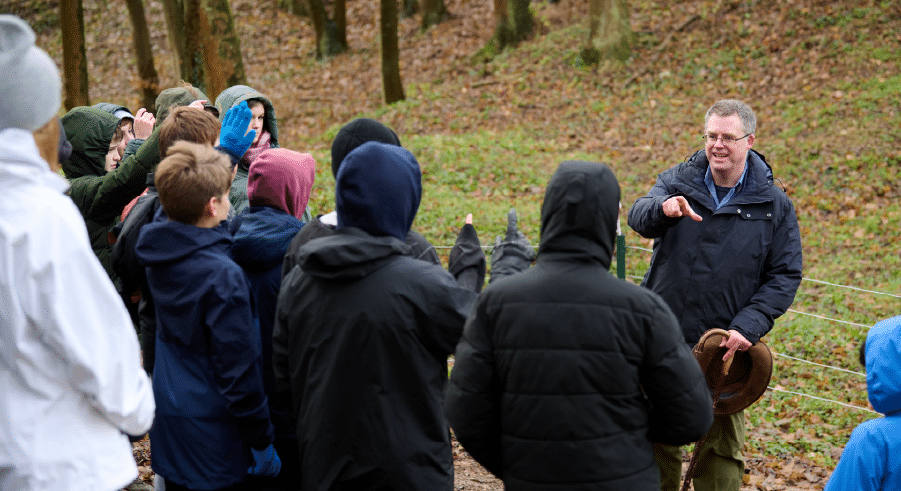 A group of people dressed in jackets listen to a man speaking outdoors in a wooded area on a cloudy day.