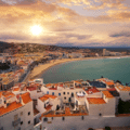 Sunset view of a coastal town with white buildings, red-tiled roofs, a curving beach, mountains in the distance, and clouds in the sky.