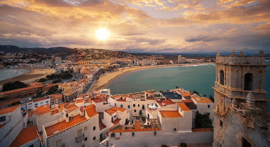 A coastal city with white buildings and red roofs overlooks a curved sandy beach and turquoise sea under a partly cloudy sky at sundown.