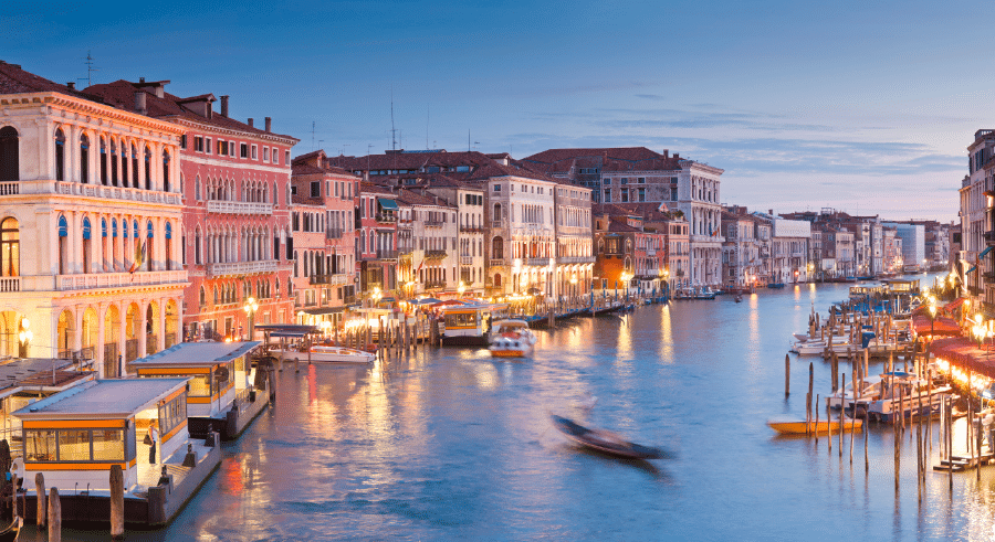 View of the Grand Canal in Venice at dusk, with historic buildings on both sides, boats on the water, and lights reflecting on the canal.
