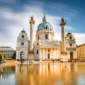 A large baroque church with two ornate columns, a central dome, and a reflecting pool in front, set under a blue sky with some clouds.