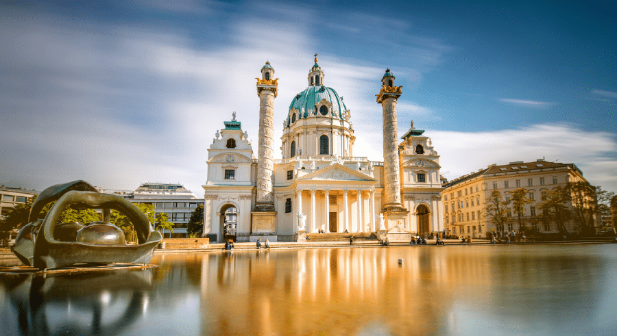 Baroque-style church with a green dome and twin columns reflected in a large pool, with modern sculpture on the left and clear blue sky above.