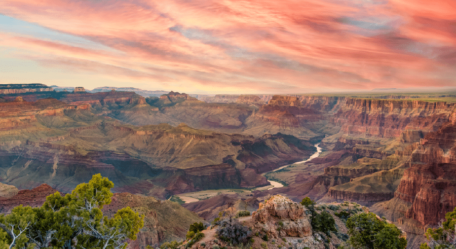 Panoramic view of the Grand Canyon at sunset with layered rock formations, the Colorado River below, and a dramatic sky with pink and orange clouds.