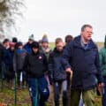 A group of people dressed in winter clothing walk in a line outdoors along a fenced path, led by a man holding a stick on a cloudy day.