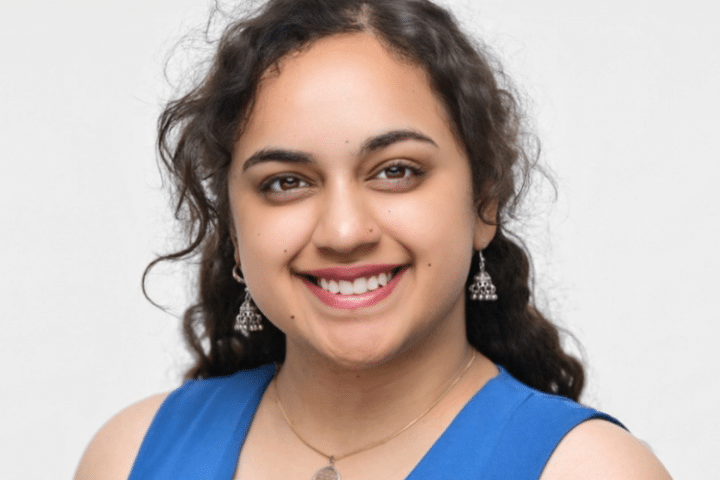 A woman with long curly hair, wearing a blue top, silver earrings, and a necklace, smiles at the camera against a plain white background.