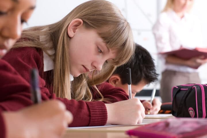 Students in a classroom, wearing red uniforms, are writing at their desks whilst a teacher stands in the background holding a book.