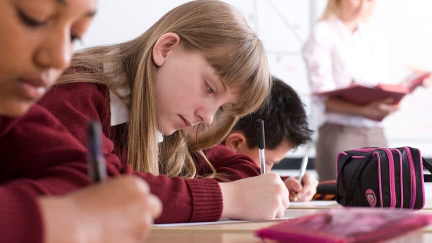 Students in a classroom, wearing red uniforms, are writing at their desks whilst a teacher stands in the background holding a book.