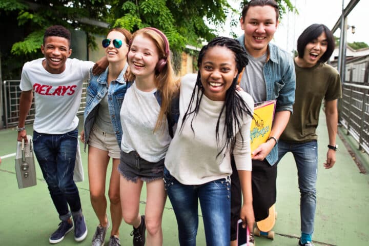 Six young adults smile and walk together outdoors on a path, with trees in the background and casual clothing, suggesting a relaxed group outing.