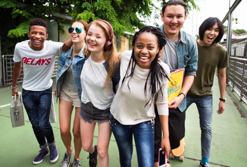 Six young adults smile and walk together outdoors on a path, with trees in the background and casual clothing, suggesting a relaxed group outing.