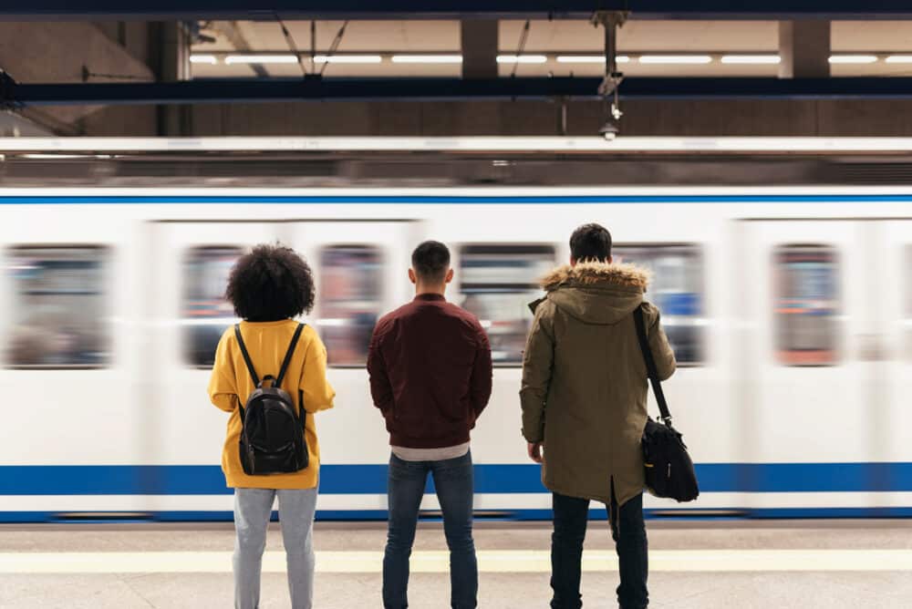 Three people with rucksacks stand on an underground platform facing a moving train, which appears blurred in motion.