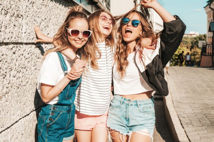 Three young women in sunglasses stand together smiling and laughing on a sunny street, wearing casual summer clothes.