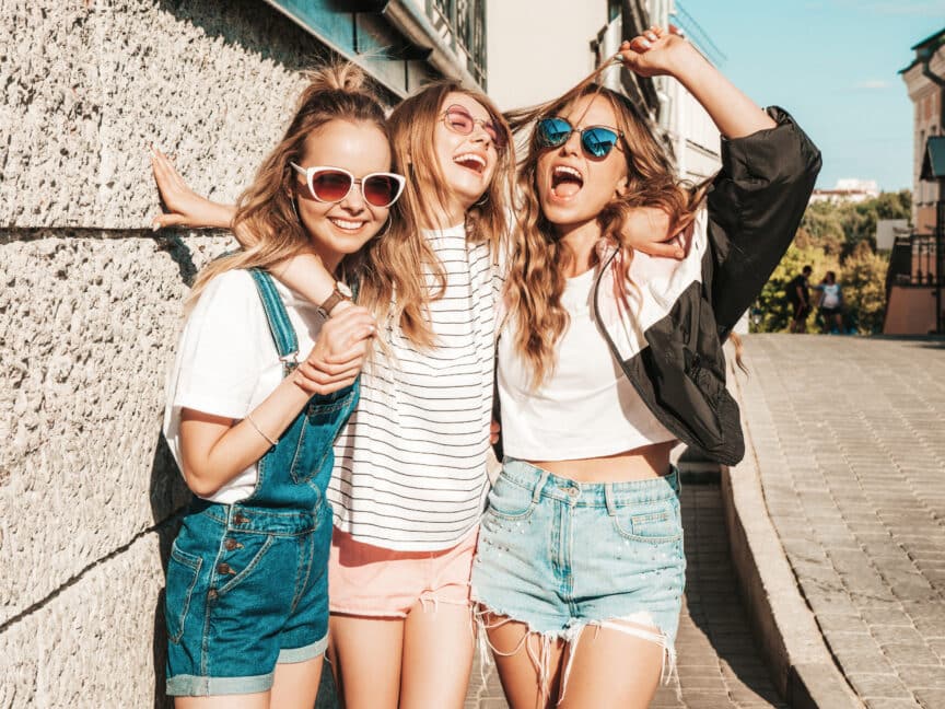 Three young women in sunglasses stand together smiling and laughing on a sunny street, wearing casual summer clothes.