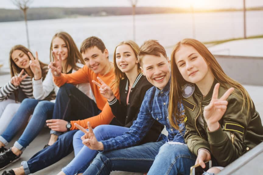 Six teenagers sit outdoors near water, smiling and making peace signs at the camera. The sun is setting in the background.