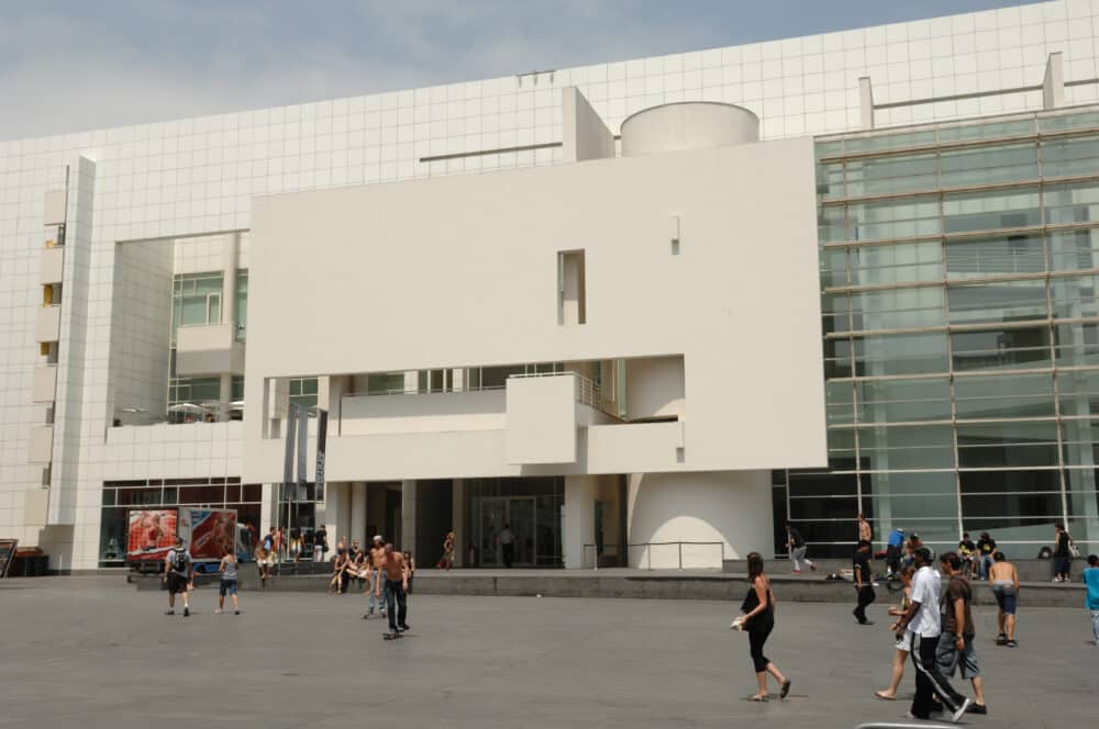 People walk and skateboard in front of a modern white building with large glass windows and geometric architectural features.
