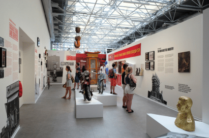 People view exhibits and sculptures in a well-lit museum gallery with informative wall displays, motorbikes, and historical artefacts.