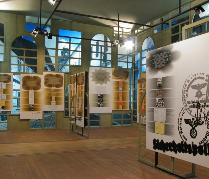 Art exhibition with modern informational panels displayed in a room with wooden floors, large windows, and track lighting.