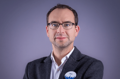Man with short brown hair, glasses, and a dark blazer stands against a plain purple background, arms folded, wearing a white shirt and a round name badge.