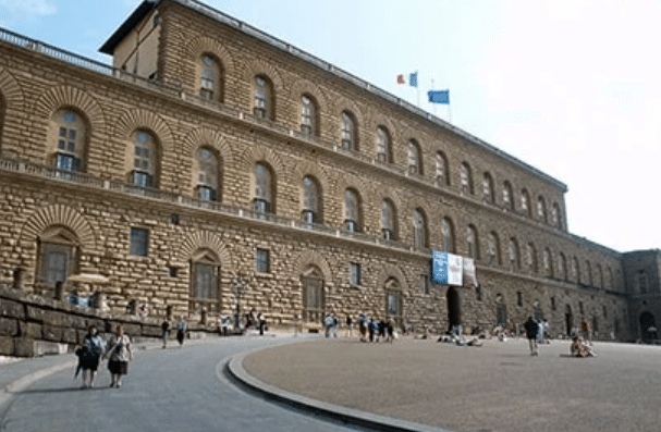 Large stone Renaissance palace with many arched windows, banners hanging above the main entrance, and people walking in a spacious courtyard.