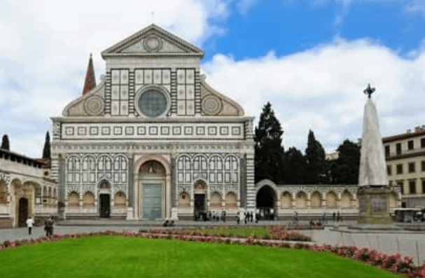 A historic church with a geometric marble façade stands in front of a manicured lawn and a stone obelisk, with people walking nearby on a cloudy day.