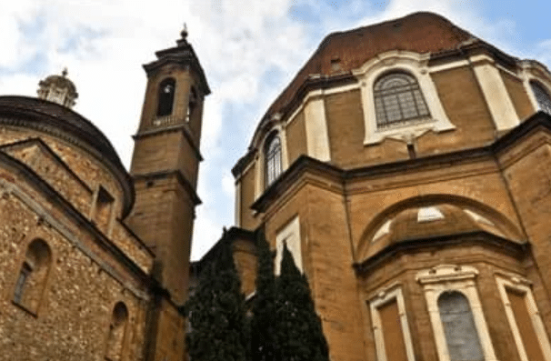 An historic church with brown brick walls, arched windows, a dome, and a bell tower, photographed from a low angle against a cloudy blue sky.